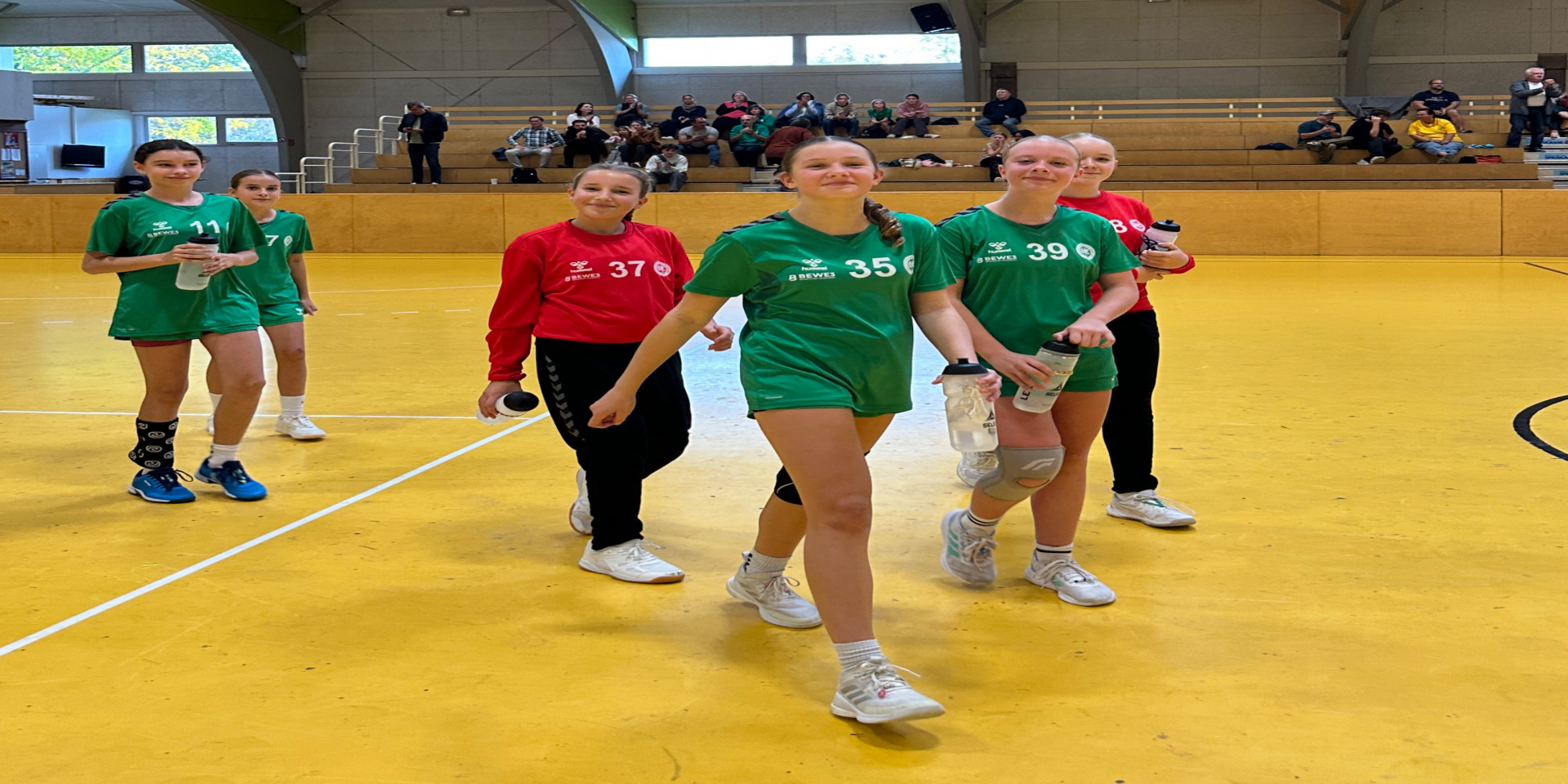Handballerinnen in grünen Trikots mit Nummern 11, 35 und 39 gehen über gelben Hallenboden.